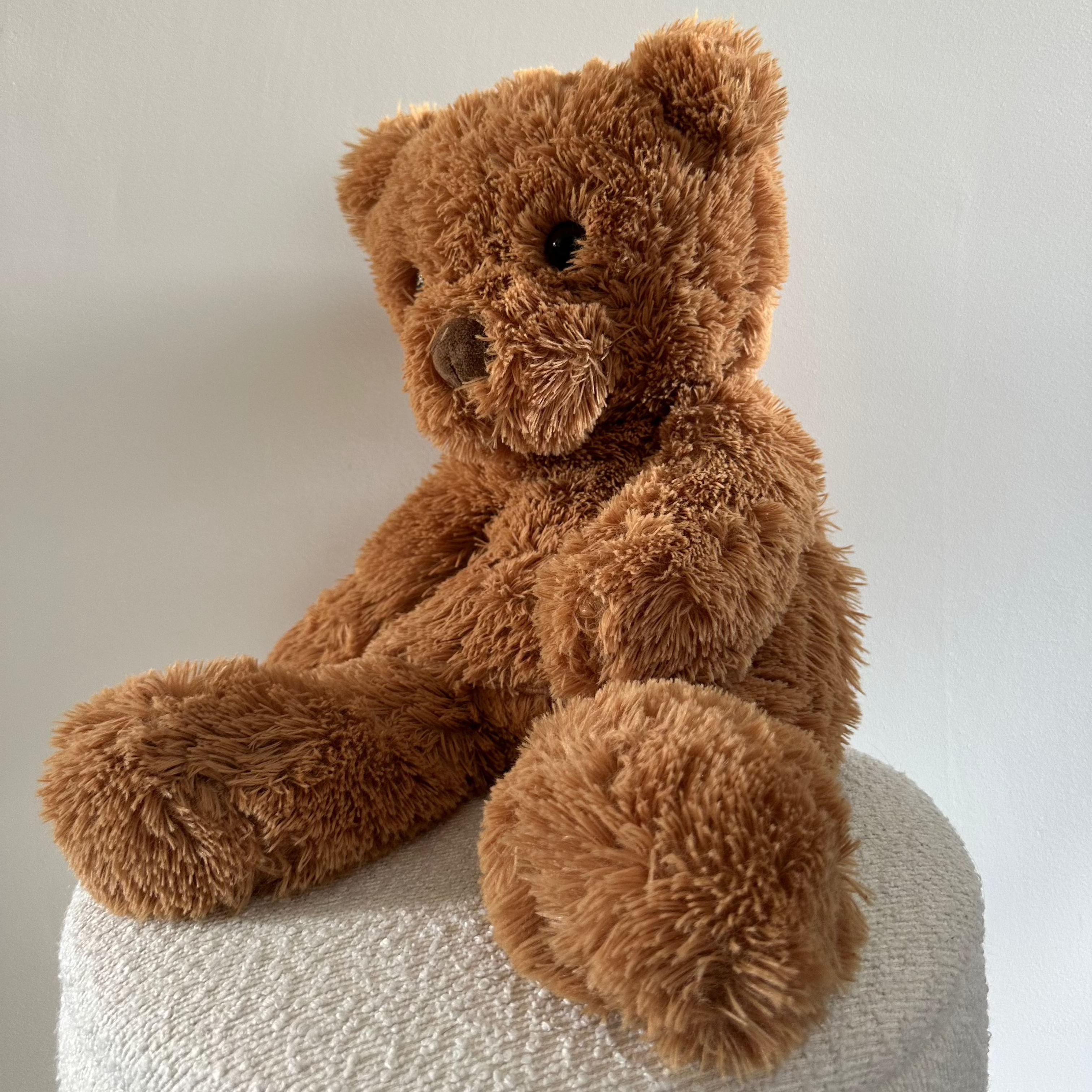 Zachte bruine verzwaarde teddybeer zittend op een witte poef tegen een lichte achtergrond.